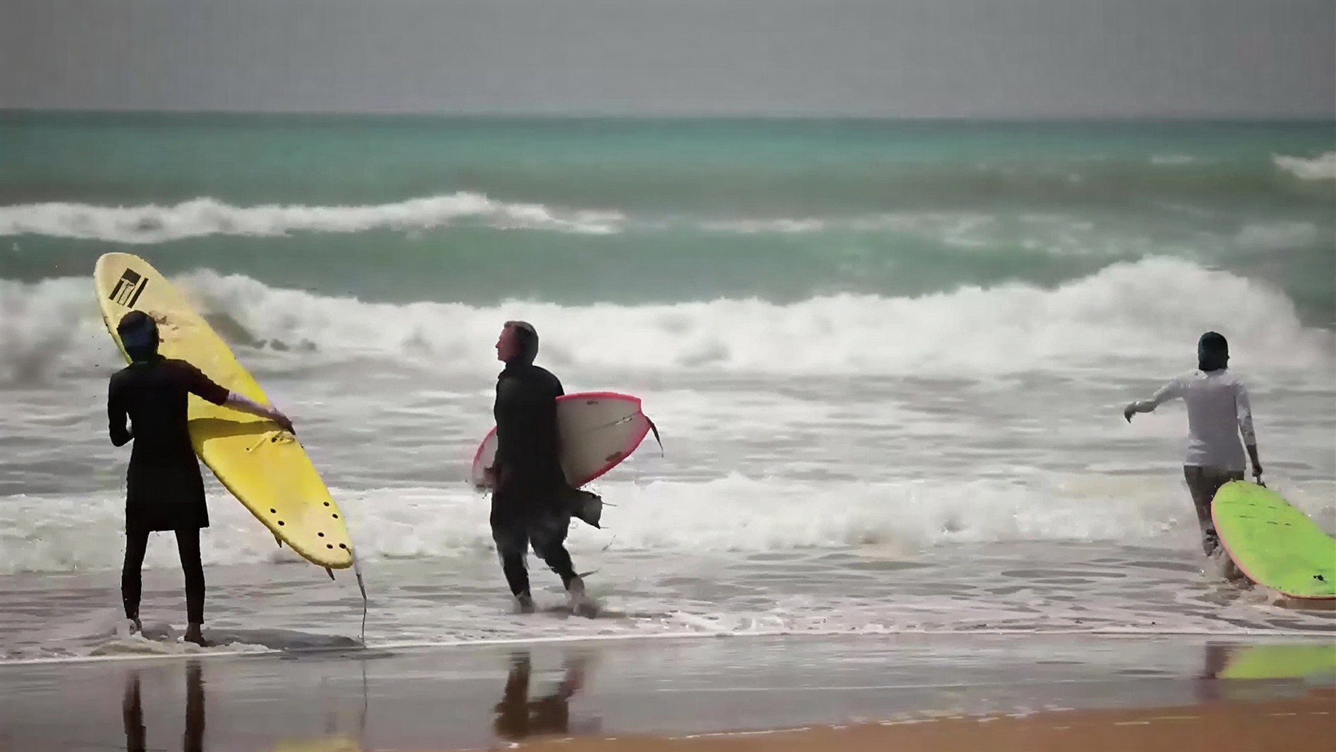 Three women in traditional clothing carrying surfboards on a sunny beach in Iran